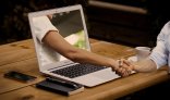 Online Das Foto zeigt eine Hand die aus einem Laptop rausschaut und schüttelt eine zweite Hand die sich vor dem Laptop befindet