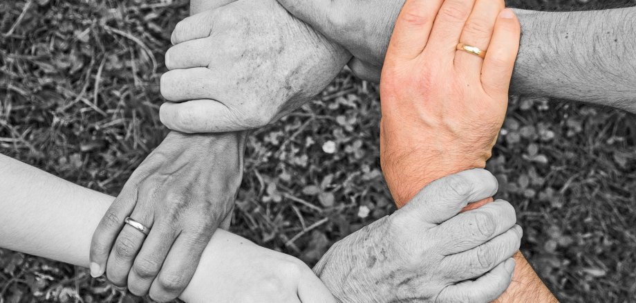 Das Foto zeigt mehrere Hände in Graustufen, die sich gegenseitig am Handgelenk halten und so einen Kreis bilden. Eine Hand ist farbig gestaltet.