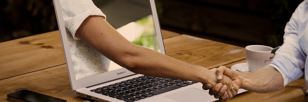 Das Foto zeigt eine Hand, die aus einem Laptop rausschaut und schüttelt eine zweite Hand, die sich vor dem Laptop befindet.
