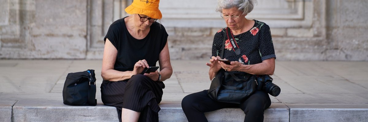 Zwei Seniorinnen sitzen auf einer Mauer und haben Smartphones in der Hand. Eine Frau hat einen Fotoapparat umhängen.